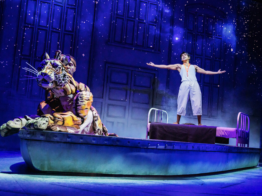 An actor stands triumphantly on a bed-turned-boat while a puppeteer manipulates a lifelike tiger puppet under a starry sky backdrop during a production of Life of Pi.