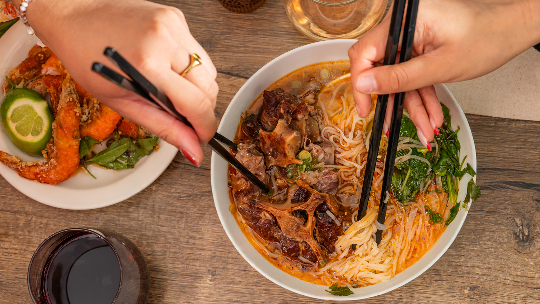 Close-up of two people using chopsticks to share a bowl of noodles topped with oxtail and fresh herbs. A bowl of crispy shrimp and a glass of wine can also be seen on the table.
