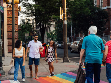 A group of people cross a colorful rainbow crosswalk in Philly's Midtown Village neighborhood, with brick buildings and leafy trees in the background.