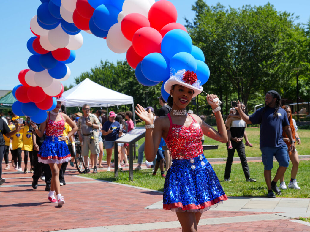 People walking in a parade line holding colorful balloons during Red, White & Blue To-Do. A woman waves at the camera