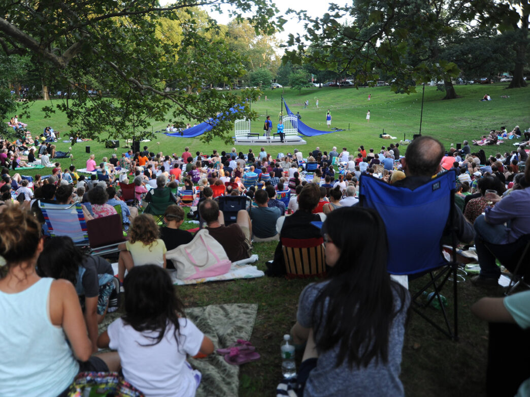 Families and friends gather on a grassy hillside under green trees at Clark Park while watching a Shakespeare performance.