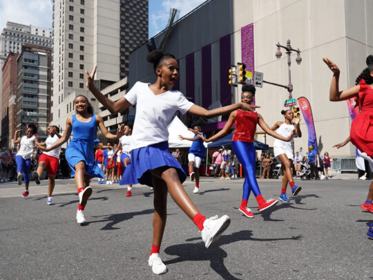 A group of young dancers in red, white and blue outfits perform a dance routine in the middle of Broad Street during the Avenue of the Arts Block party. Philadelphia's City Hall is visible in the background.