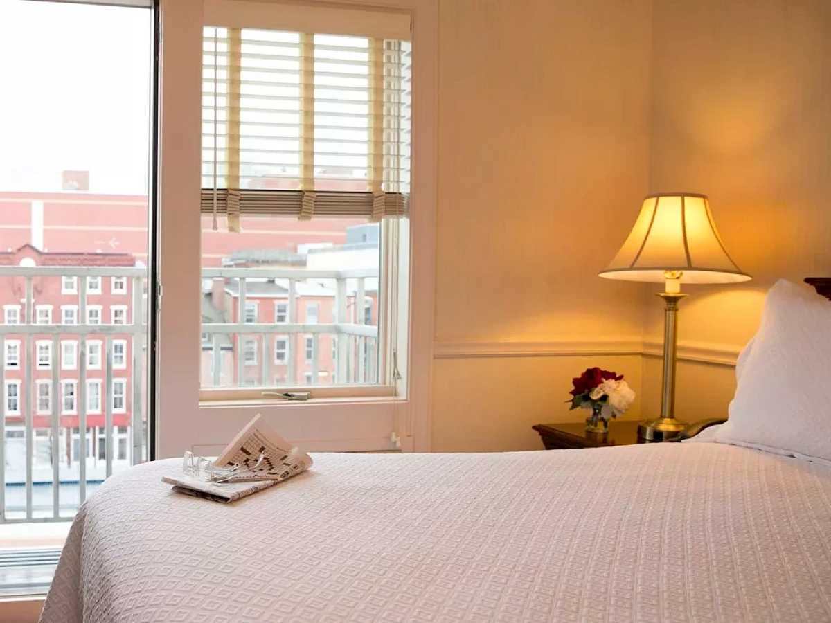 A cozy guest room with a a bed with white linens and a bedside table with a lamp and flower. Large windows offer a view of Old City's historic buildings.
