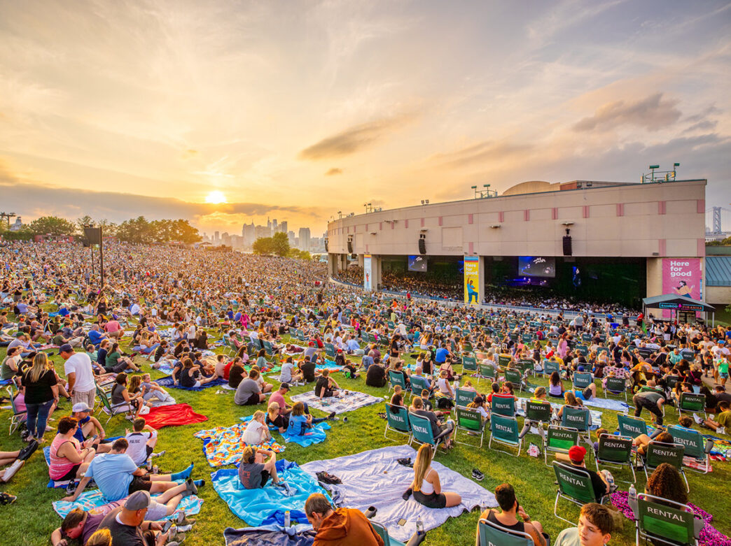 Crowds of people sitting on blankets and lawn chairs fill a large grassy hillside facing the Freedom Mortgage Pavilion during a concert. The Philadelphia skyline and setting sun is seen in the distance.