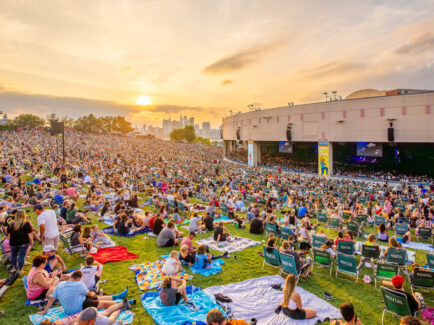 Crowds of people sitting on blankets and lawn chairs fill a large grassy hillside facing the Freedom Mortgage Pavilion during a concert. The Philadelphia skyline and setting sun is seen in the distance.