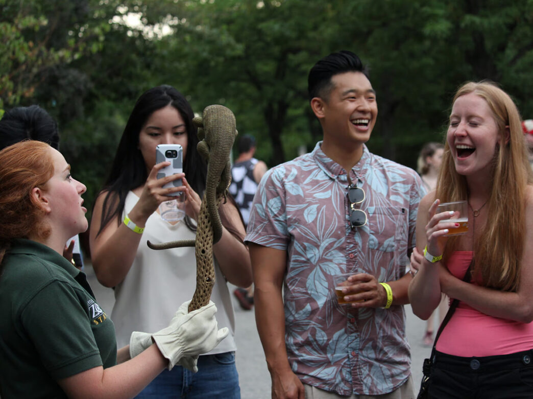 A group of smiling young adults holding cups of beer interact with a zookeeper holding a lizard at the Philadelphia Zoo's Ale Festival.