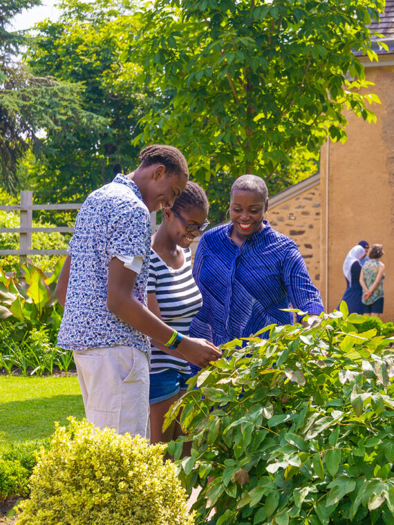 A group of people explore a lush garden on a sunny day at Bartram's Garden, with historic stone buildings in the background.