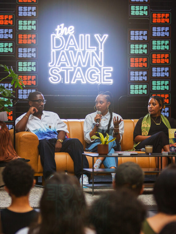 A group of panelists speak on stage at BlackStar Film Festival's Daily Jawn Stage in front of a live audience, framed by plants and vibrant neon sign.