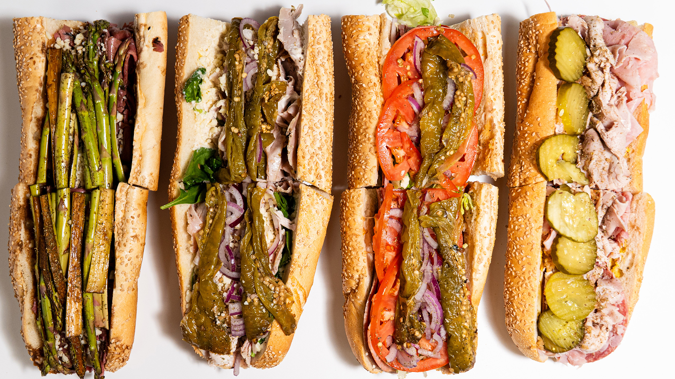 An overhead view of four hoagies from Fink's Hoagies, each filled with a variety of meats, cheeses, vegetables and toppings on sesame seed rolls.