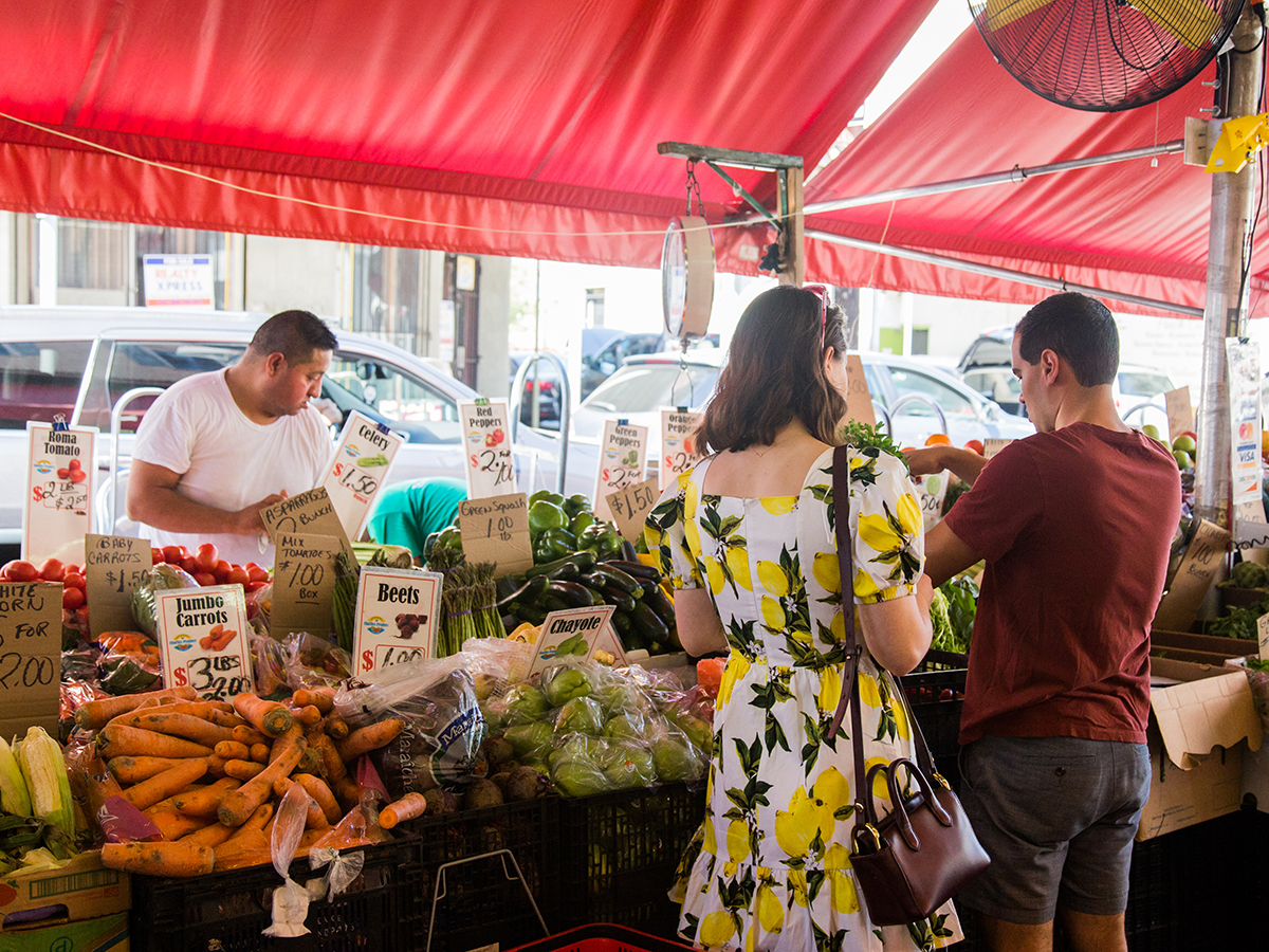 Shoppers browse fresh produce at a vendor stand under a bright red awning at the South 9th Street Italian Market.