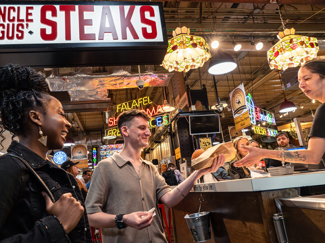 A cheesesteak in a brown paper bag is handed off to a customer at the counter of Uncle Gus' Steaks in Reading Terminal Market.