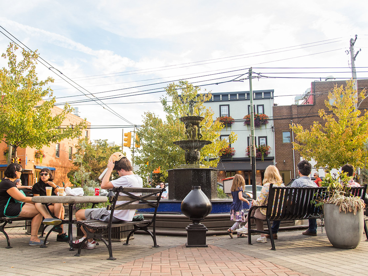 People sit and relax on benches around the Singing Fountain in East Passyunk, with autumn trees and charming storefronts in the background.