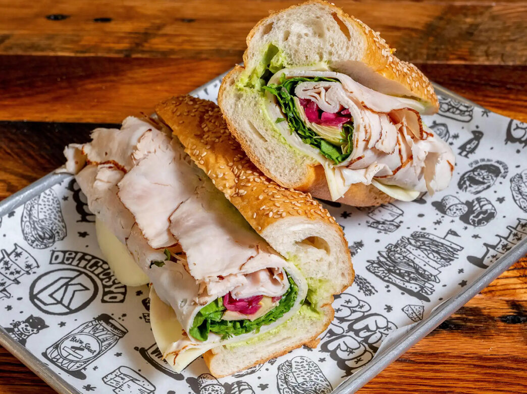 A sesame seed hoagie roll stuffed with turkey, pesto, cheese, lettuce and more on a tray lined with Liberty Kitchen branded paper.